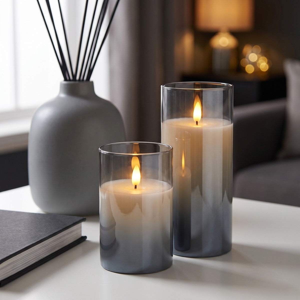 Two lit VEKE candles in smoky gray on a white table beside a grey vase